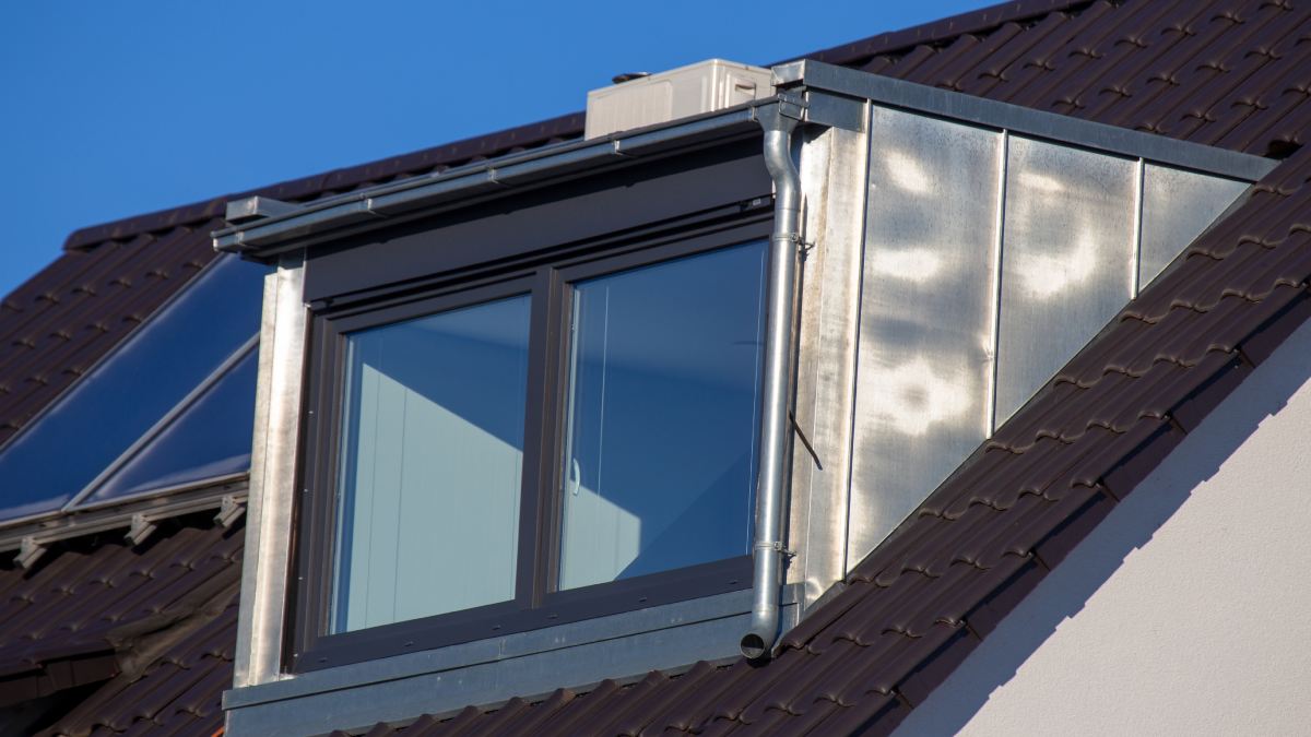 Lucarne de toit en zinc sur toit en tuiles foncées.