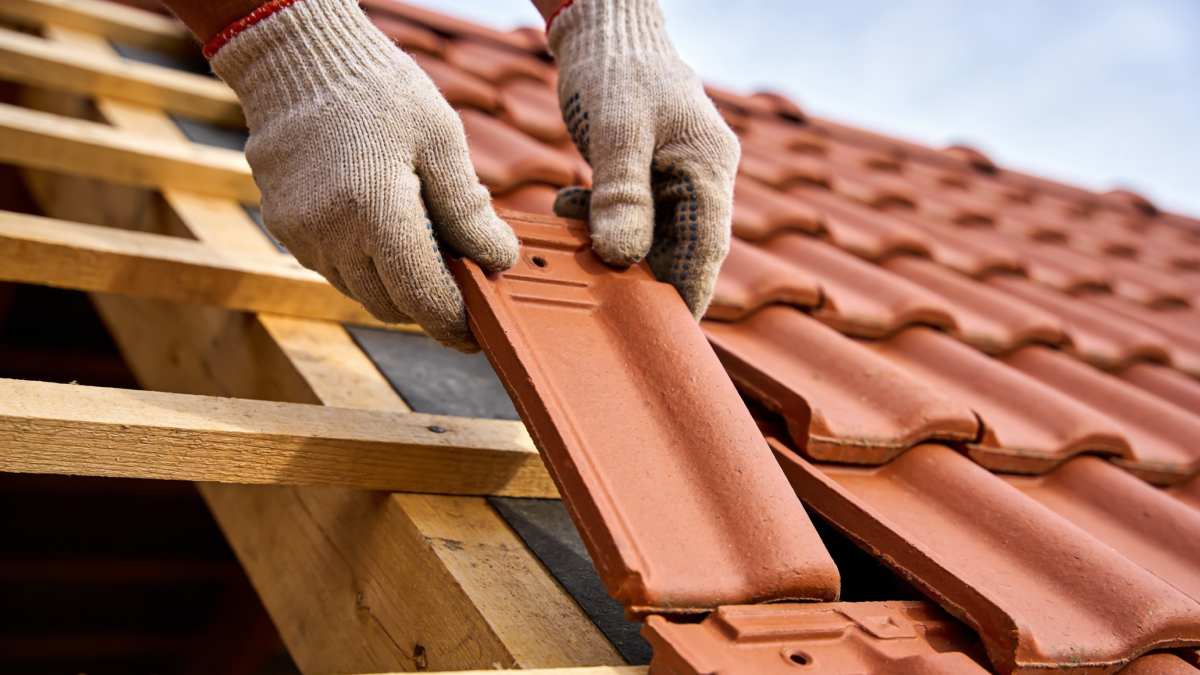 Un gros plan montre des mains gantées plaçant soigneusement un carreau d'argile rouge sur une structure de toit, soulignant le processus de construction et de réparation.
