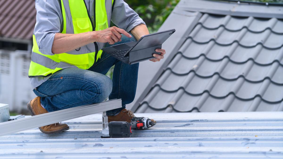 Un travailleur de la construction vêtu d'un gilet haute visibilité s'accroupit sur un toit métallique tout en utilisant une tablette, avec une perceuse électrique à proximité.