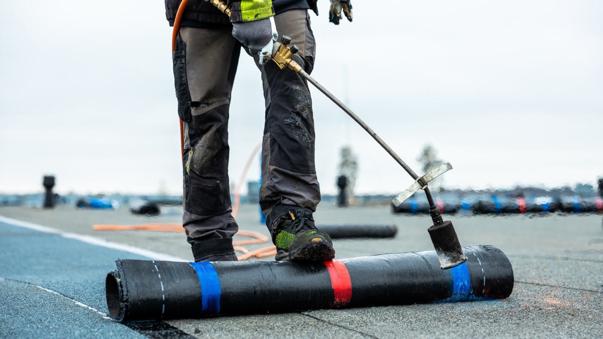 Un travailleur en équipement de protection utilise une torche au propane pour appliquer une membrane de toiture noire sur un toit plat, avec des matériaux supplémentaires et un ciel nuageux visible.