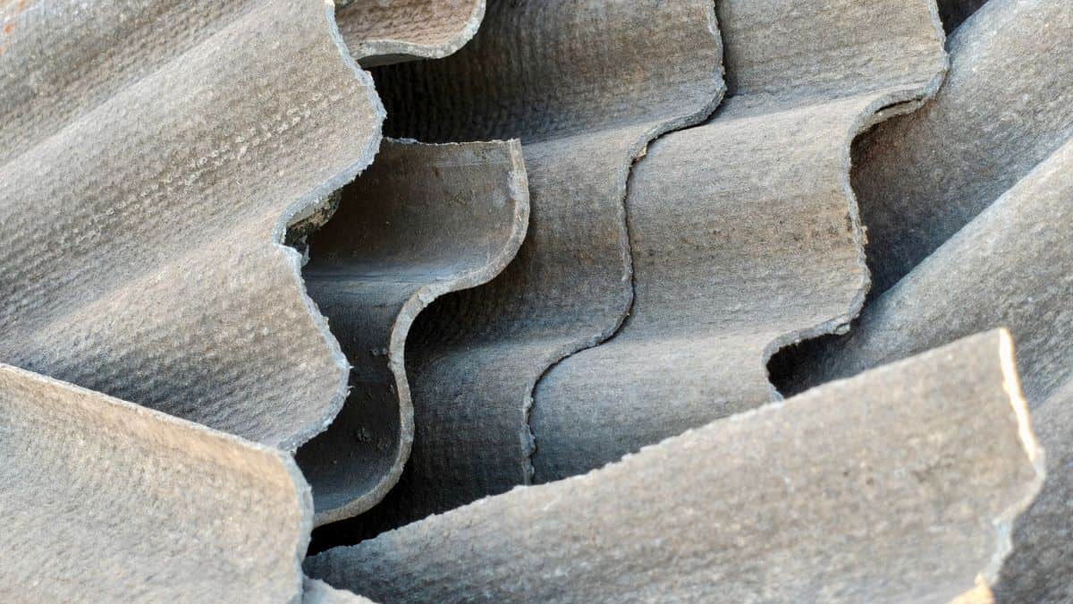 Gros plan sur de vieilles feuilles de toiture en amiante-ciment ondulé brisées à texture grise altérée, montrant des fissures dangereuses et des bords déchiquetés par l'âge et la détérioration.
 